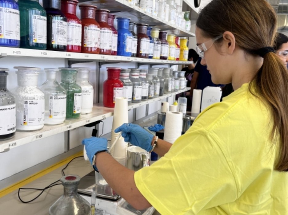 Schülerin vor einem Gestell Laborflaschen, gefüllt mit farbigen Pigmenten, am Abwiegen von Pulver.