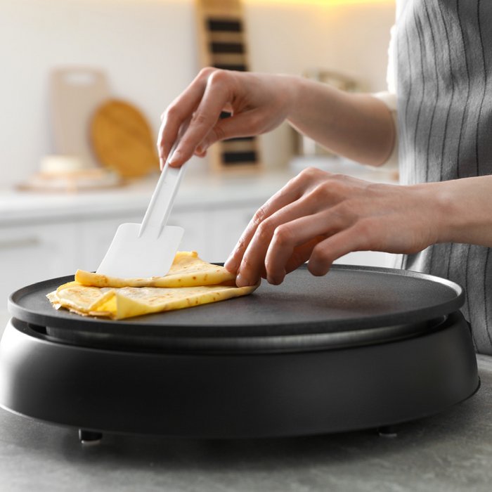 Eine Crêpe wird auf einer schwarz beschichteten Crêpesplatte gebraten und mit einem weissen Teigschaber gefaltet