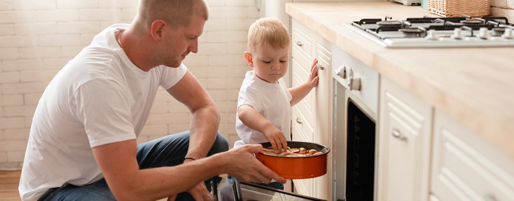 Young dad with a toddler is pushing an apple cake in a orange spring form into the oven.