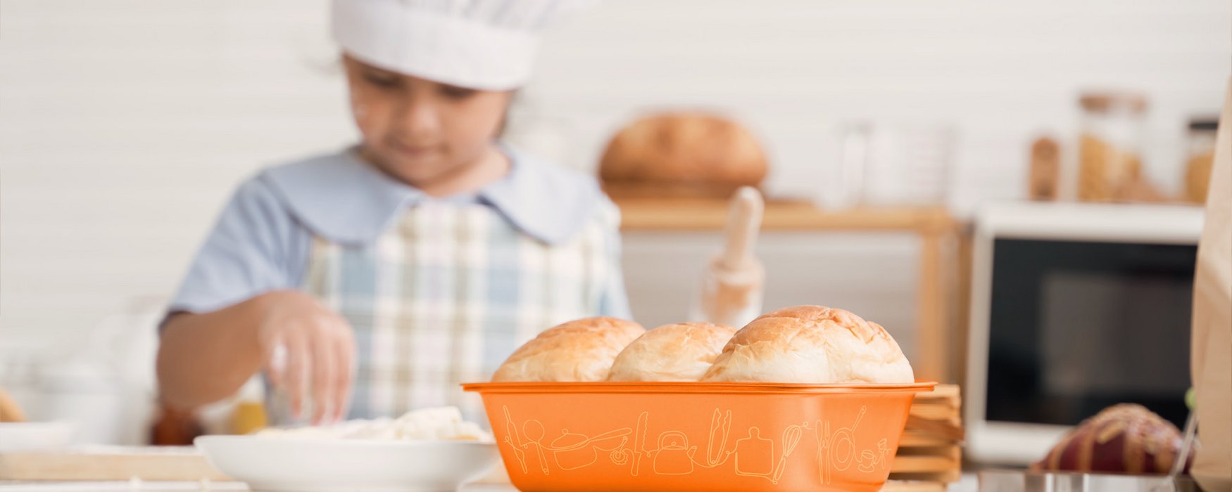 Im Vordergrund eine orange Brot-Ofenform mit aufgedruckten Motiven. Im Hintergrund ein kleines Mädchen mit Kochmütze