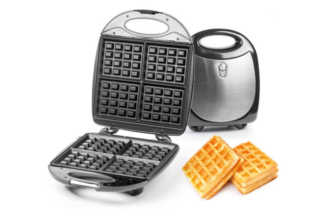 Ein silbernes Waffeleiesen steht geschlossen im Hintergrund und ein offenes mit schwarzer Innenbeschichtung im Vordergrund. Daneben liegen gebackene Waffeln.
