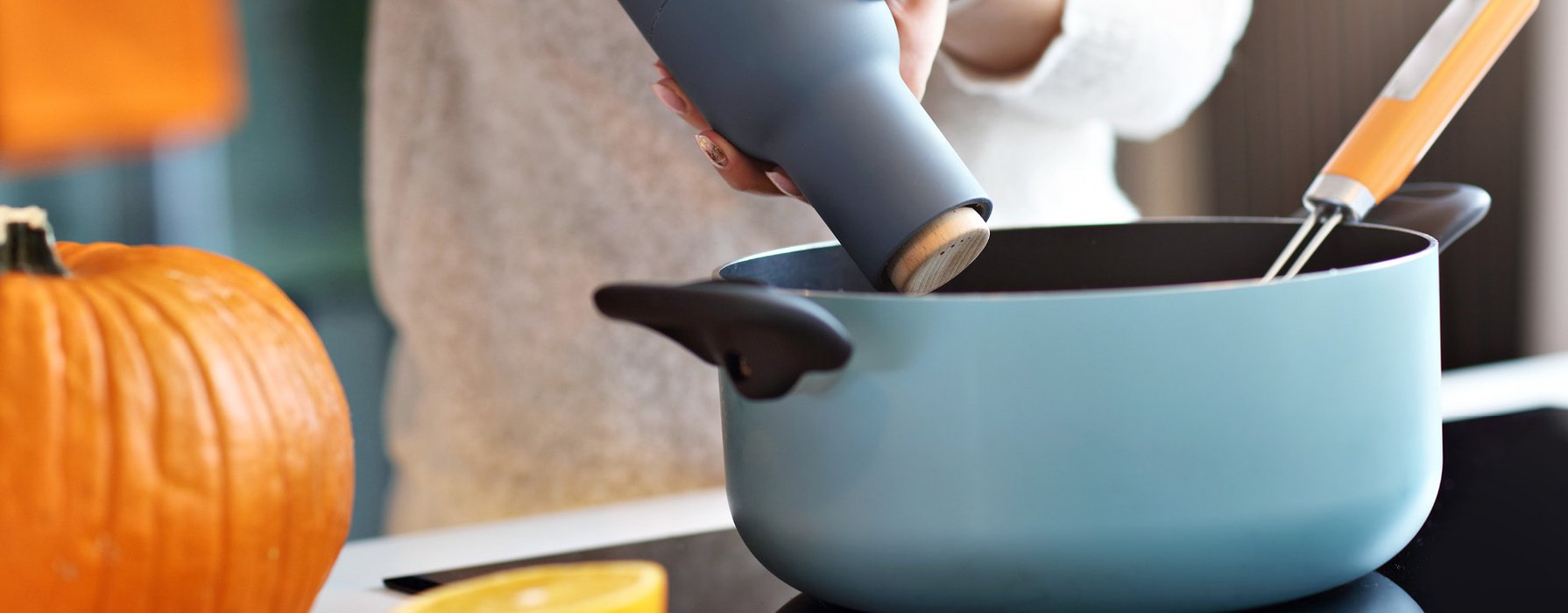 A pot with a light blue coated outside and a black coating on the inside is standing on a stove while somebody is seasoning with a pepper mill.