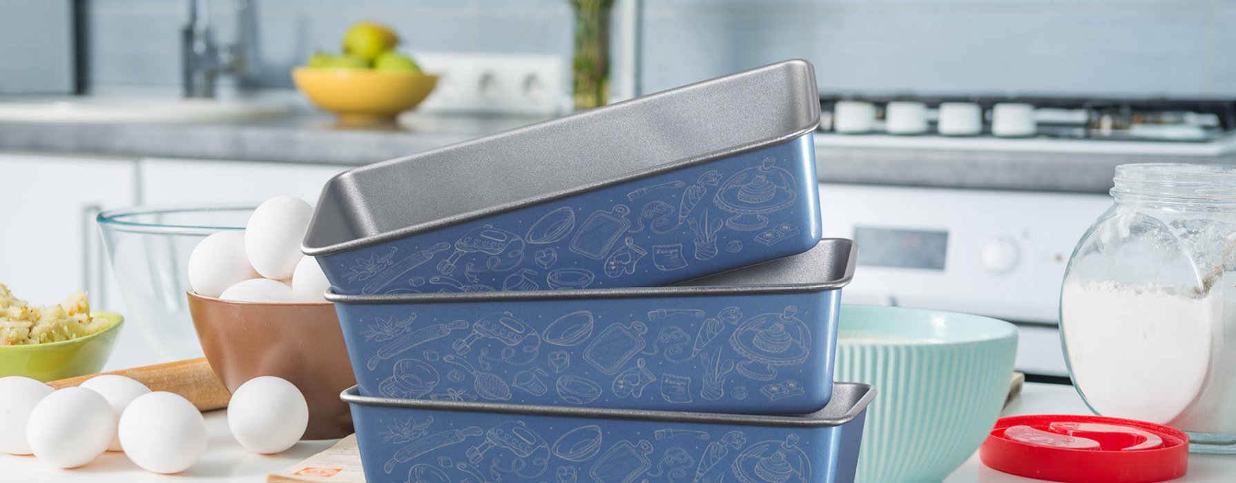 Three stacked loaf pans with blue coating on the outside and silver ornaments printed on it are standing with baking ingredients on a countertop