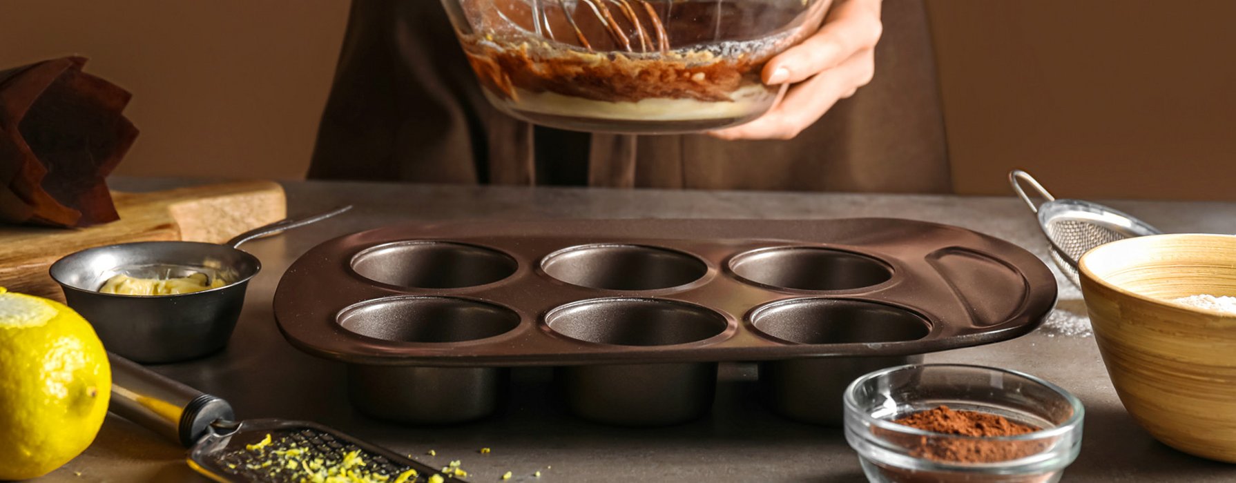 There is a black coated muffin pan on the table in which chocolate dough is filled in.