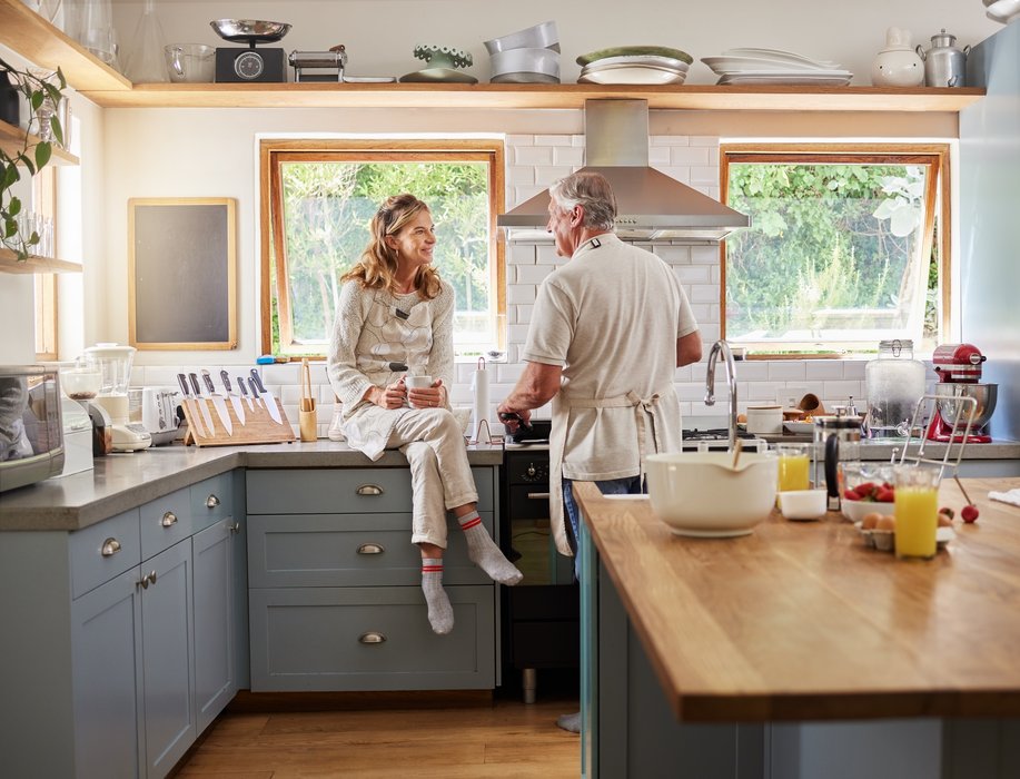 A man is cooking a meal in a big kitchen. Next to him a woman is sitting on the counter with a cup of tea and talks to the man. 