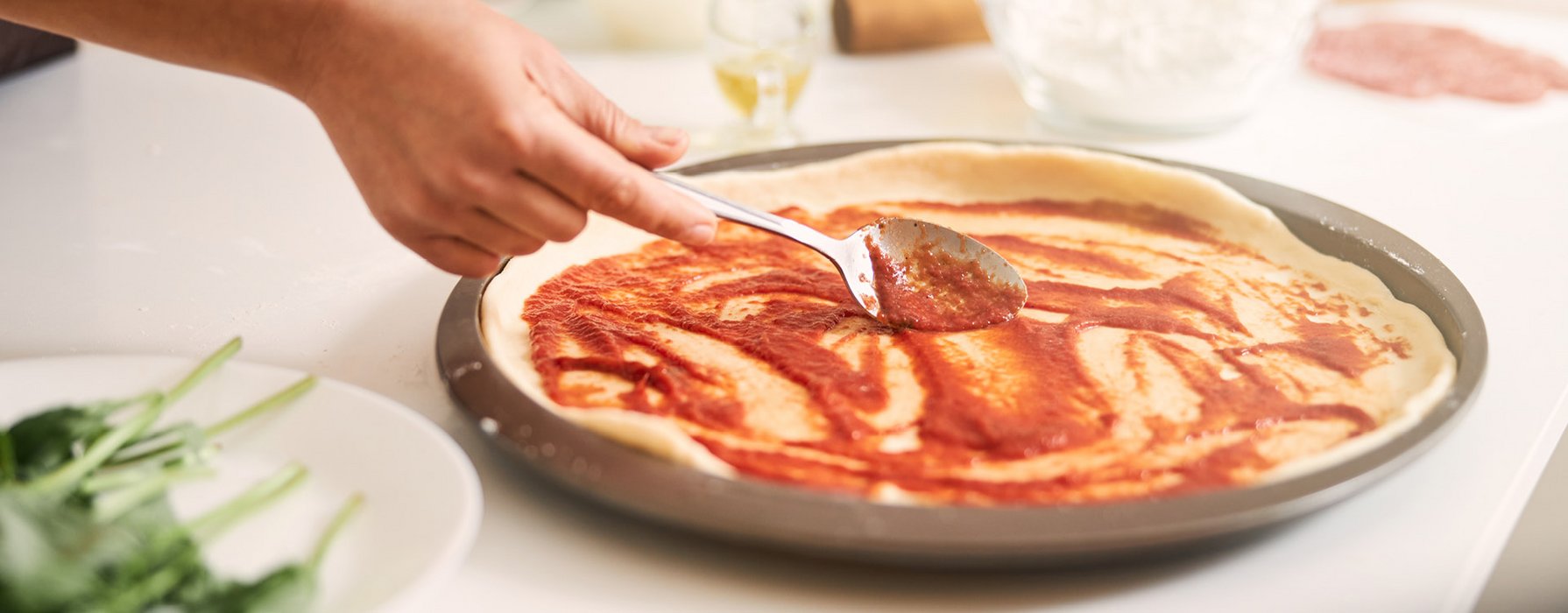 Auf einem Pizzateig, der in einem beschichteten Blech liegt, wird mit einem Löffel Tomatensauce verteilt