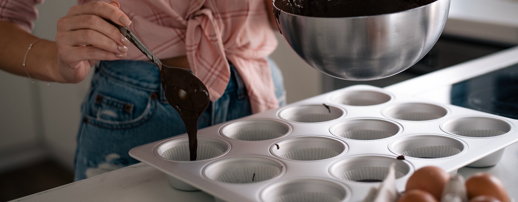 A woman is filling some dark chocolate dough with a spoon into a silver muffin tray