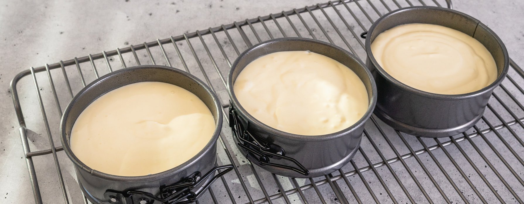 Three anthracite colored springform pans filled with dough are standing next to each other on a baking grid