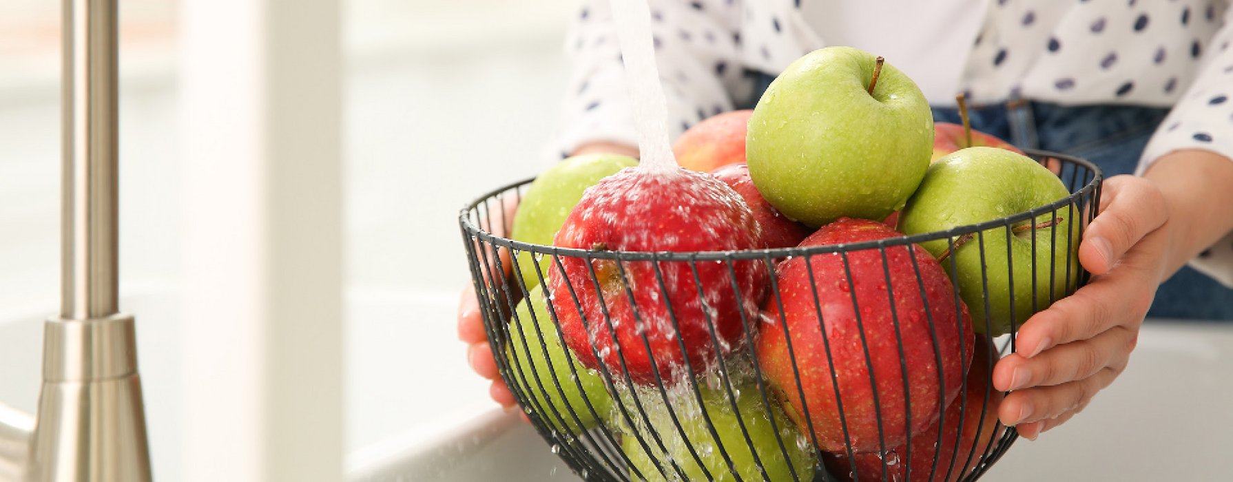 A black coated grid basket for fruits with red and green apples in it is holding under the water of a tap
