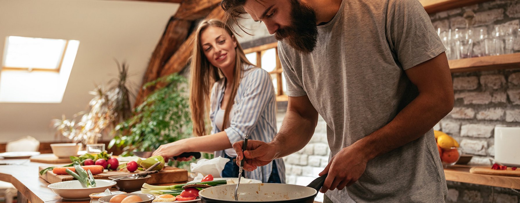 Junges Paar bereitet ein Essen in einer rustikalen Küche zu. Die Frau schneidet Gemüse, der Mann brät in einer Bratpfanne Fleisch an